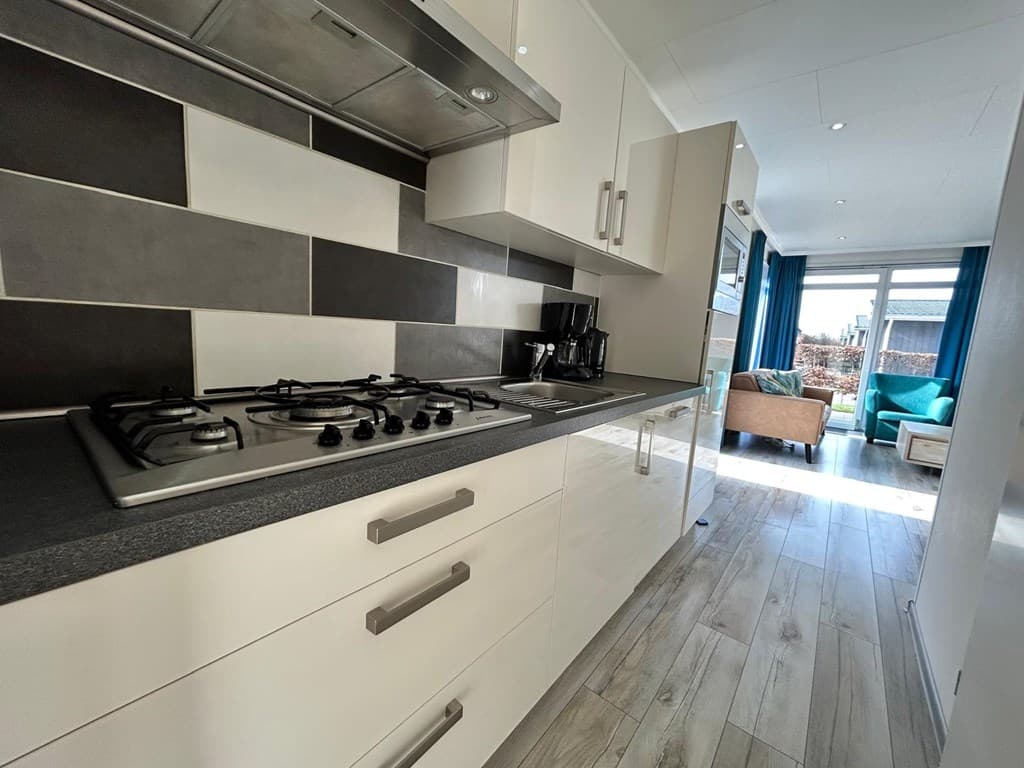 Moderne keuken met gasfornuis, zwarte en witte tegels, en toegang tot een lichte woonkamer met raam en zithoek.