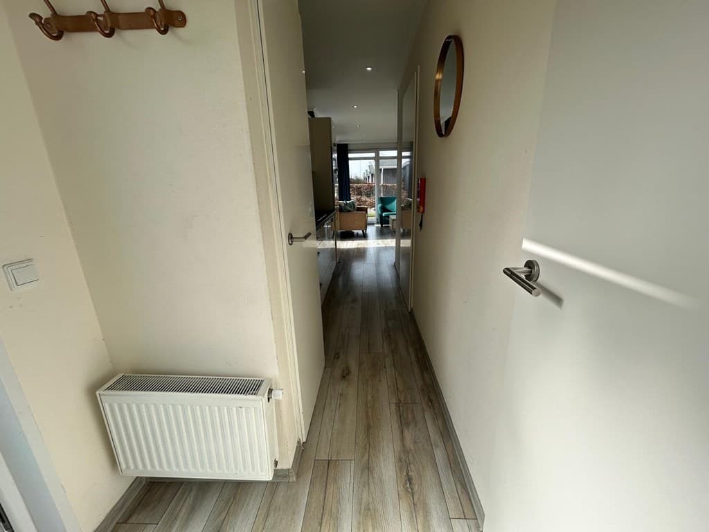 Lange gang met laminaatvloer, wandplank en ronde spiegel, leidt naar een lichte woonkamer en kitchenette.
