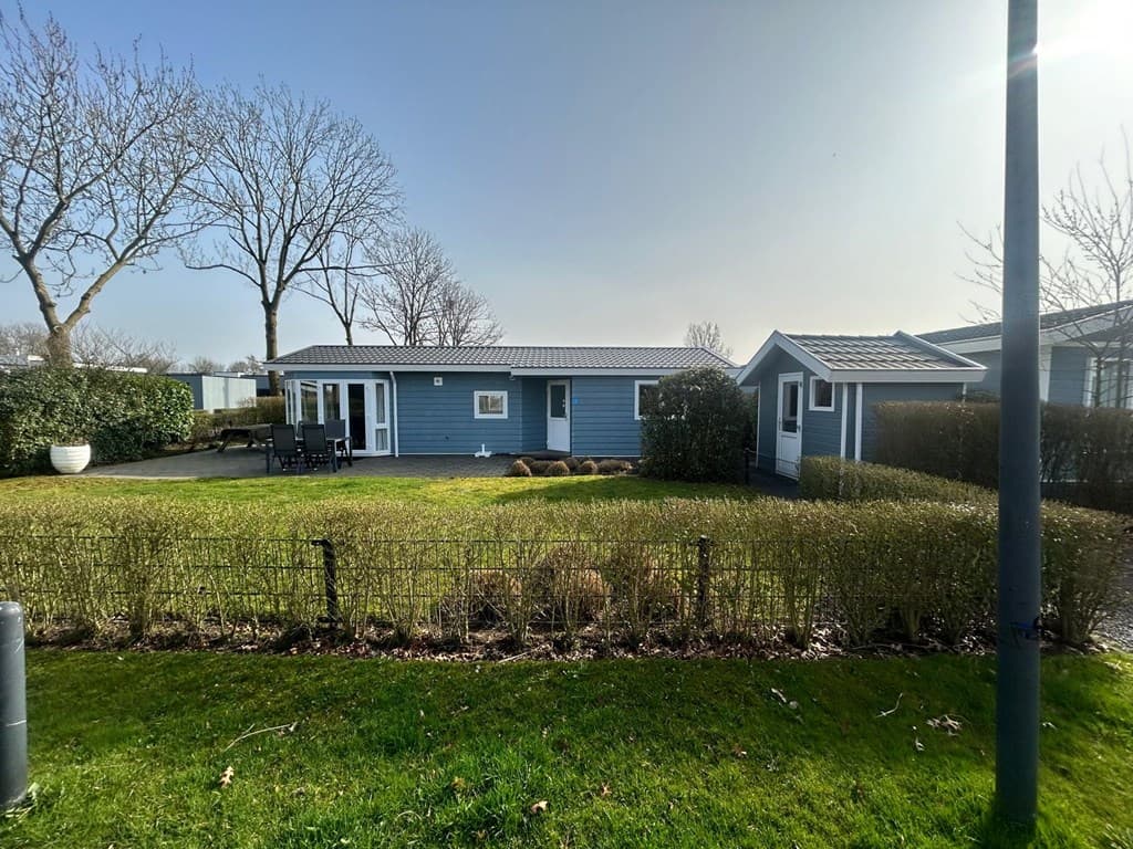 Buitenzicht op een blauwe vakantiewoning met terras en een kleine aangebouwde opbergruimte, omgeven door groene hagen.