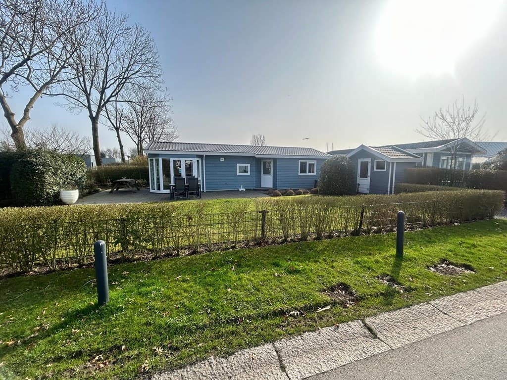 Blauwe vakantiewoning met terras, omringd door groene hagen en bomen, gelegen aan een rustige straat