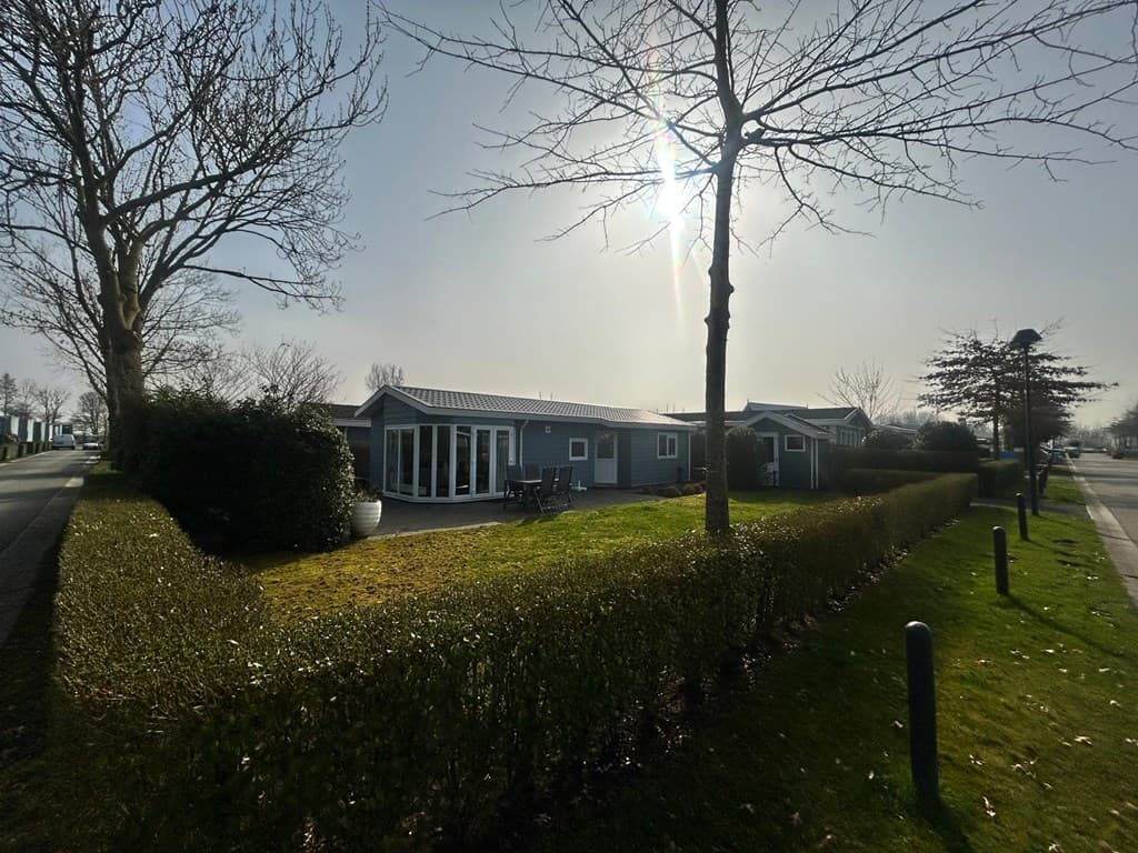 Buitenaanzicht van een vakantiewoning met groene tuin en terras, omringd door lage heggen en bomen, onder een zonnige lucht
