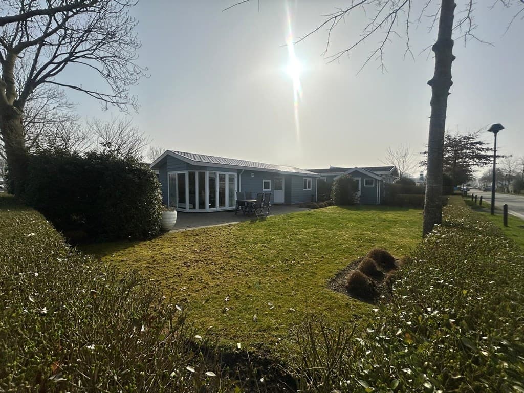 Buitenzijde van een chalet met een omheinde tuin en terras, tegen een lichte hemel met de zon erbij