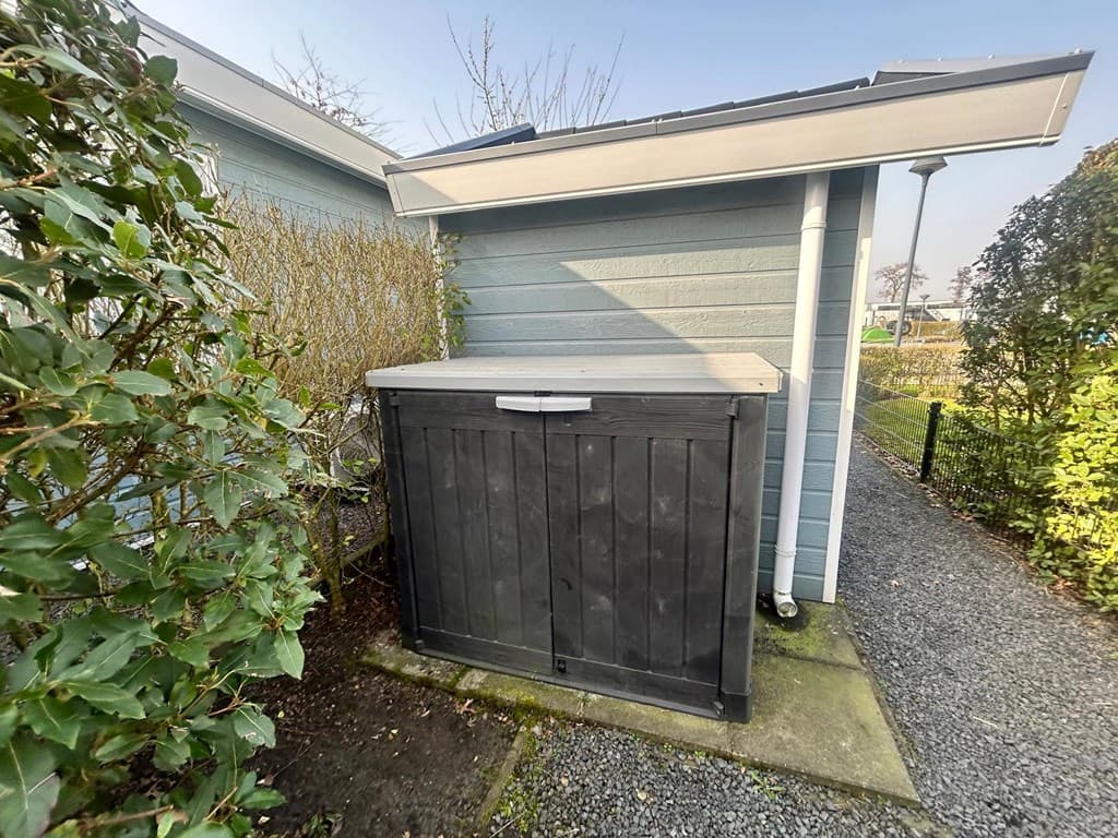 Zwarte houten berging naast een woning met blauwe gevel en grindpad in de tuin rondom.