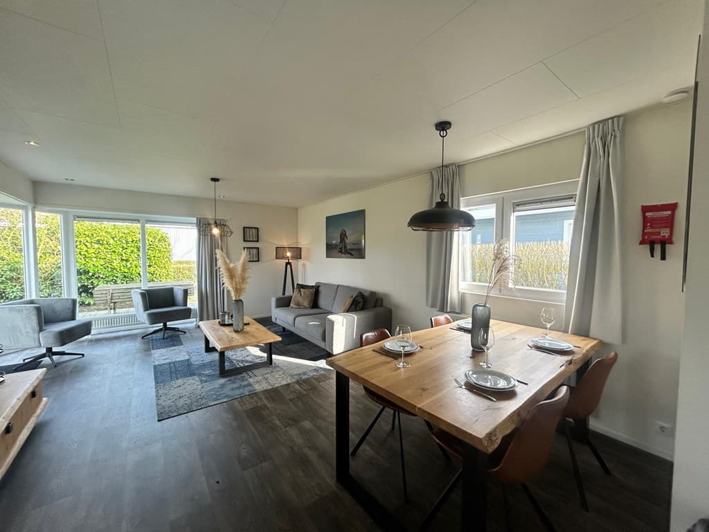 Woonkamer met eetgedeelte, bank, grote ramen en een vloerkleed in een moderne vakantiewoning