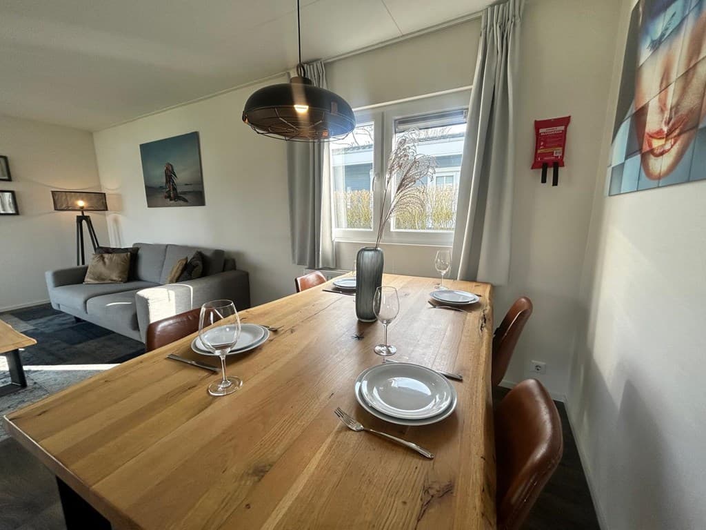 Eetruimte met houten tafel, stoelen, bord en bestek, naast een zithoek met een bank en wanddecoratie.