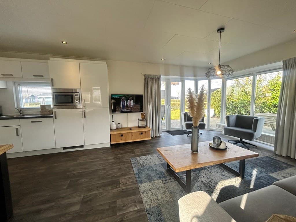Lichte woonkamer met open keuken, TV, eettafel en toegang tot een tuin met groen uitzicht.
