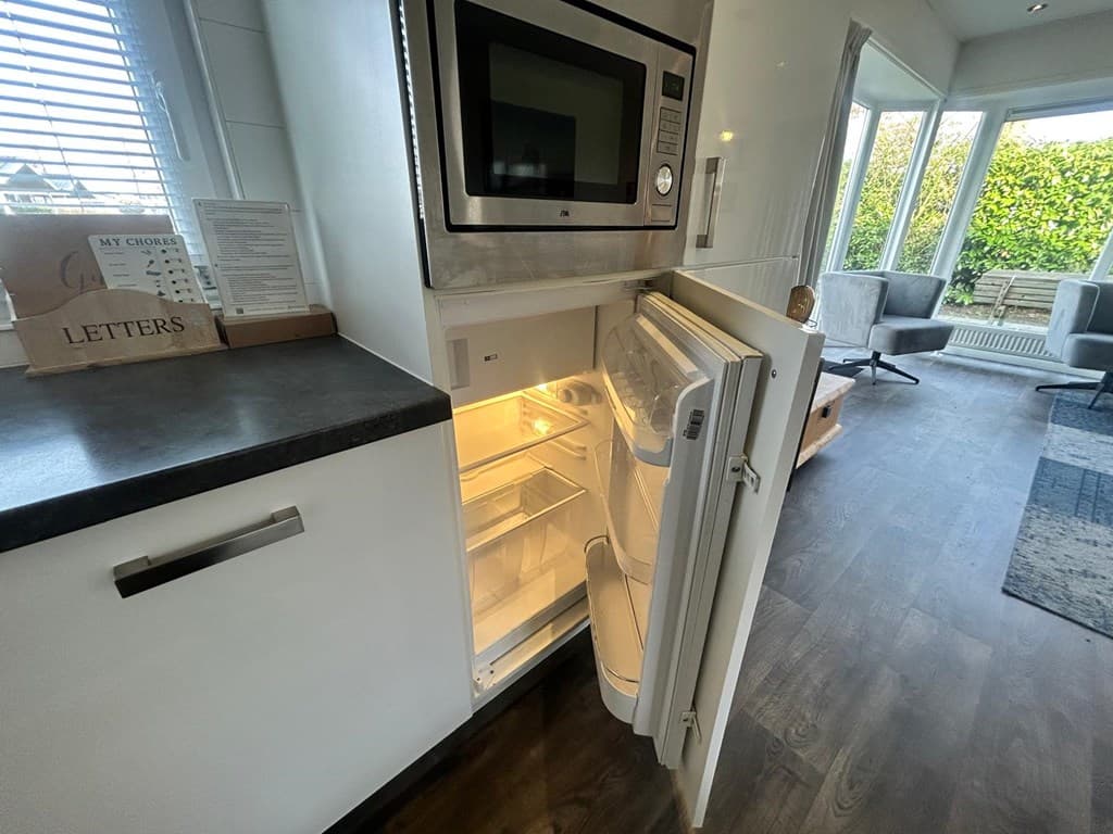 De foto toont een open koelkast met een magnetron erboven, zichtbaar in een moderne woonkamer met uitzicht op de tuin.