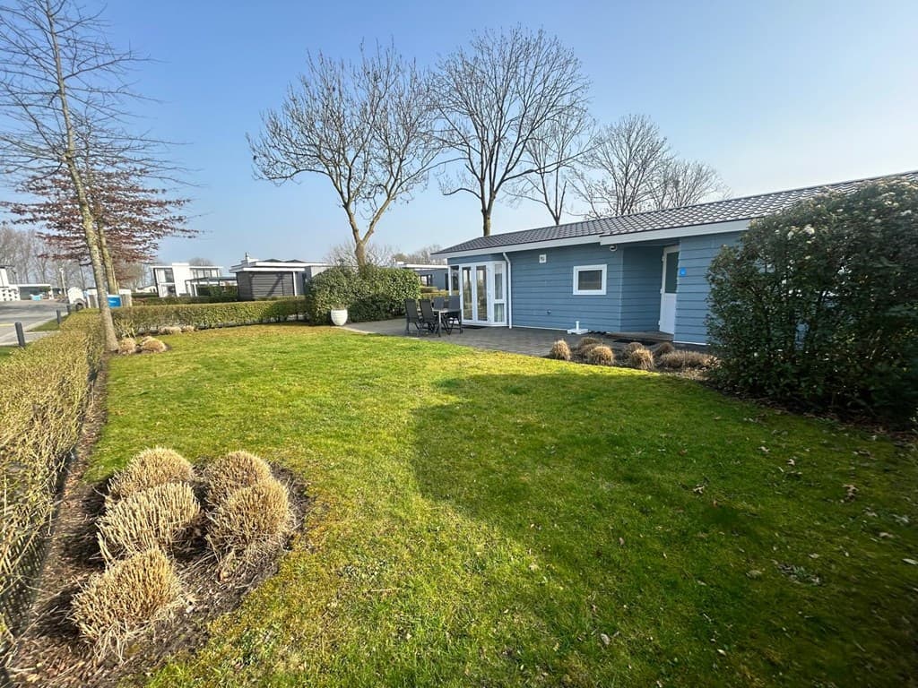Groene tuin met geschoren hagen, gras, en een terras met stoelen bij een blauw houten huisje in een rustige omgeving