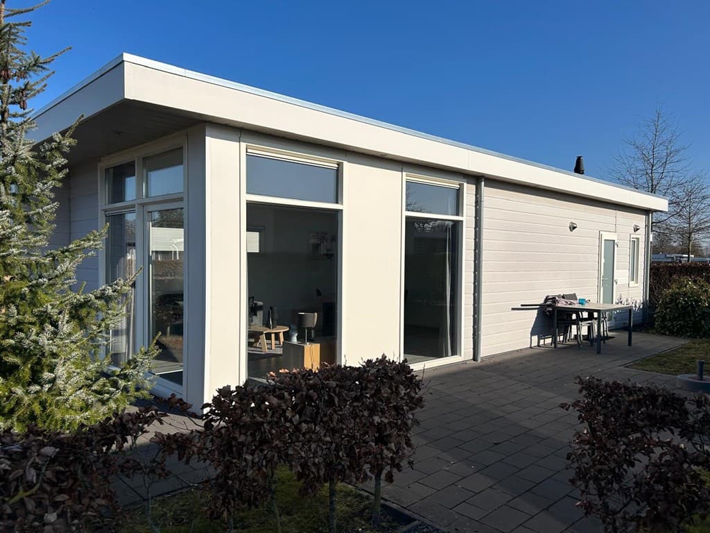 Moderne vakantiewoning met grote ramen en een terras, omgeven door struiken en een groene omgeving.