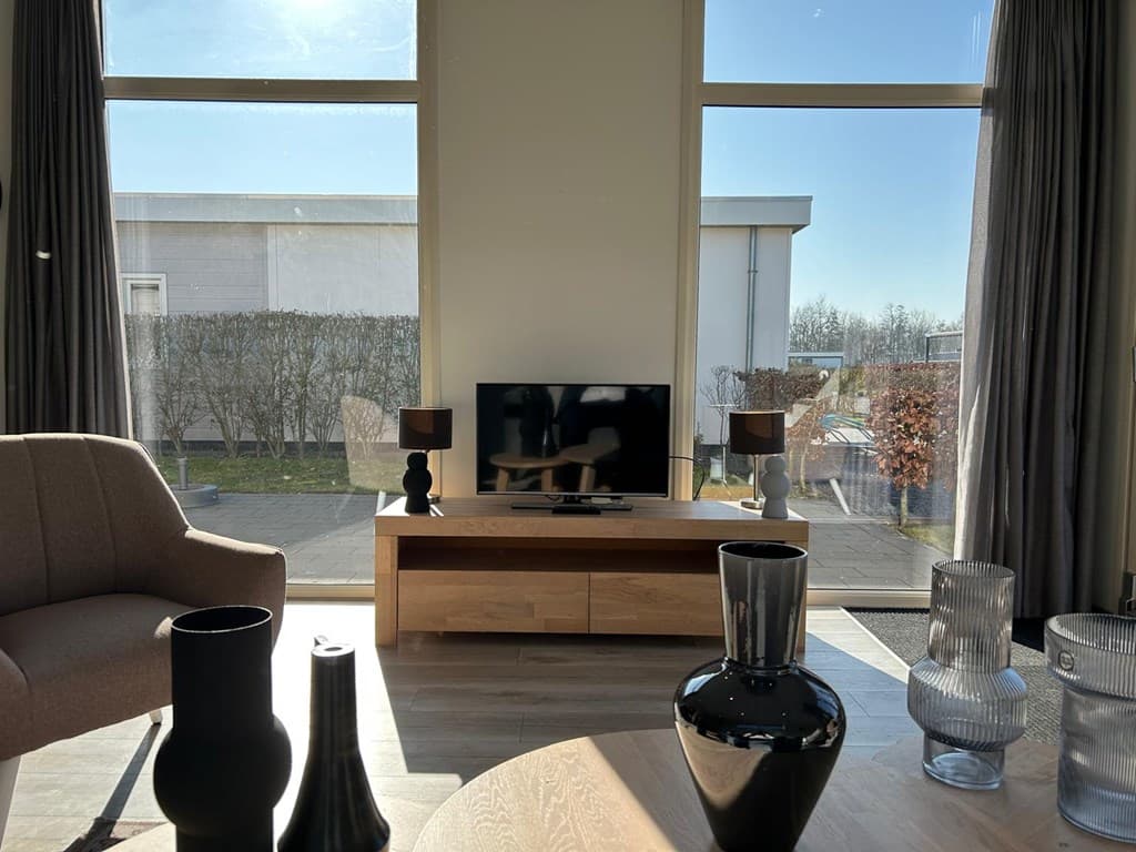 Lichte woonkamer met grote ramen, tv op een houten tv-meubel en moderne decoratie zoals vazen en lampjes.