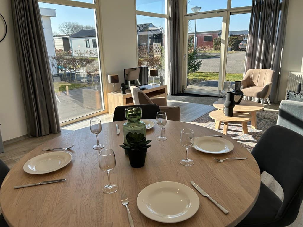 Eethoek met een ronde tafel, glazen en borden, uitzicht op buiten en een zithoek met fauteuils en een tv