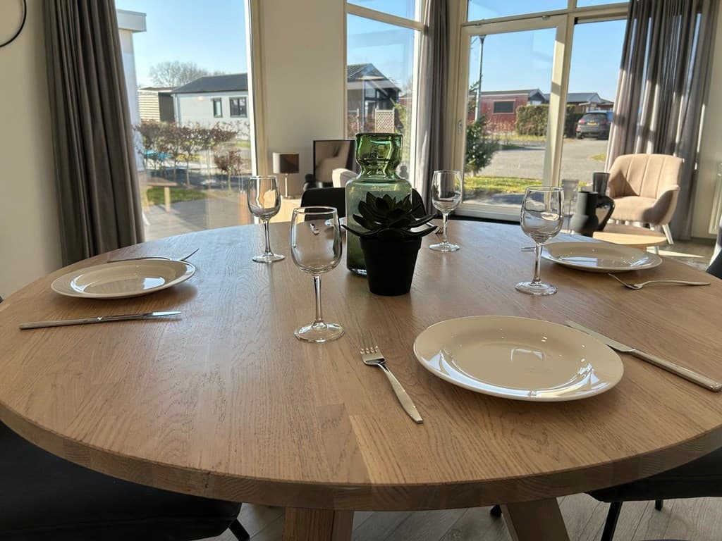 Eettafel met vier borden, glazen en een groene vaas in een lichte ruimte met grote ramen en uitzicht op buiten.
