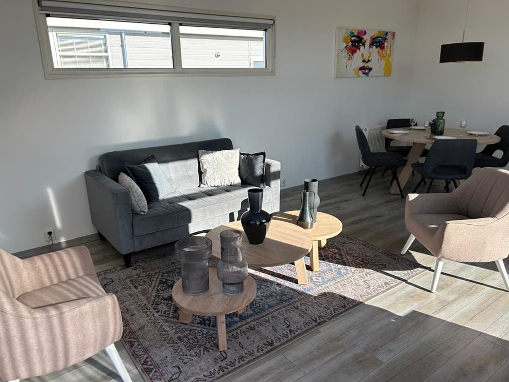 Zitgedeelte met een grijze bank, houten tafels, stijlvolle stoelen en kunst aan de muur in een lichte ruimte.
