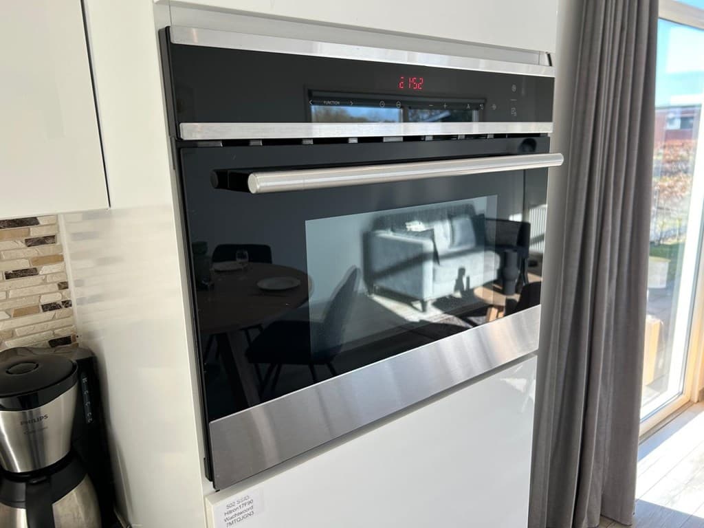 Inbouwoven met glazen deur en metalen handgreep, zicht op de eetruimte en grote ramen op de achtergrond.