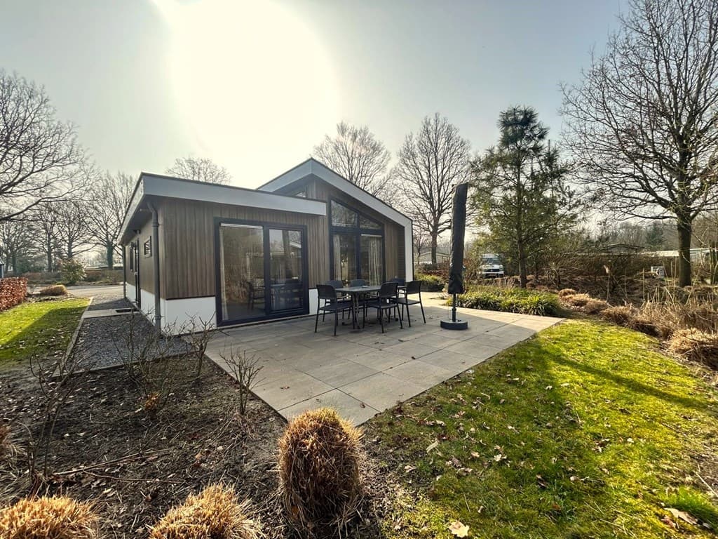 Moderne vakantiewoning met grote ramen, terras en tuin, omringd door bomen en struiken.