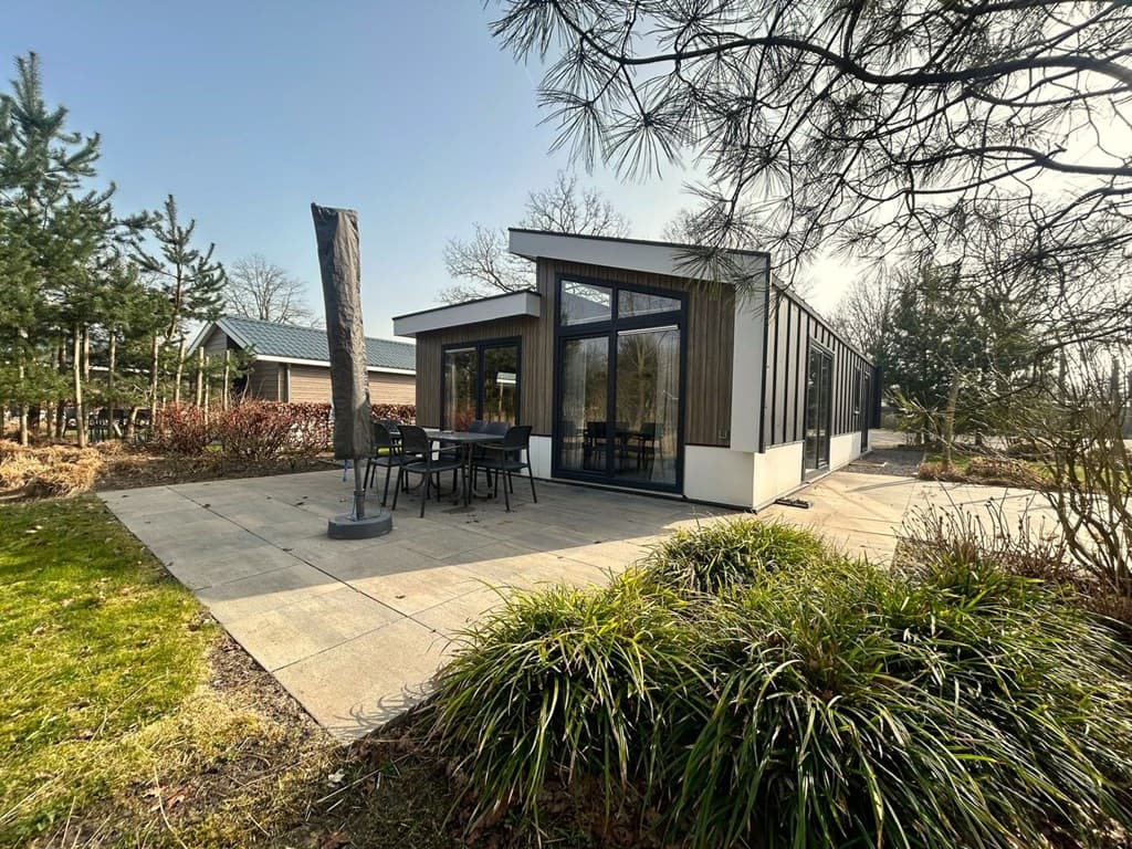 Moderne vakantiewoning met grote ramen, terras en tuinmeubilair op een betegende buitenruimte omgeven door groen.