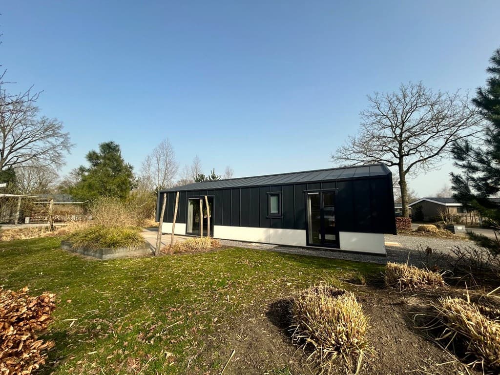 Moderne vakantiewoning met zwart-witte gevel, gelegen in een groene omgeving met bomen en gras.