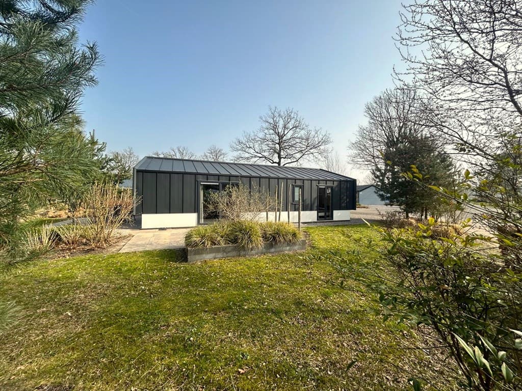 Moderne vakantiewoning met zwarte gevel en grote ramen, omringd door groen en bestrating.
