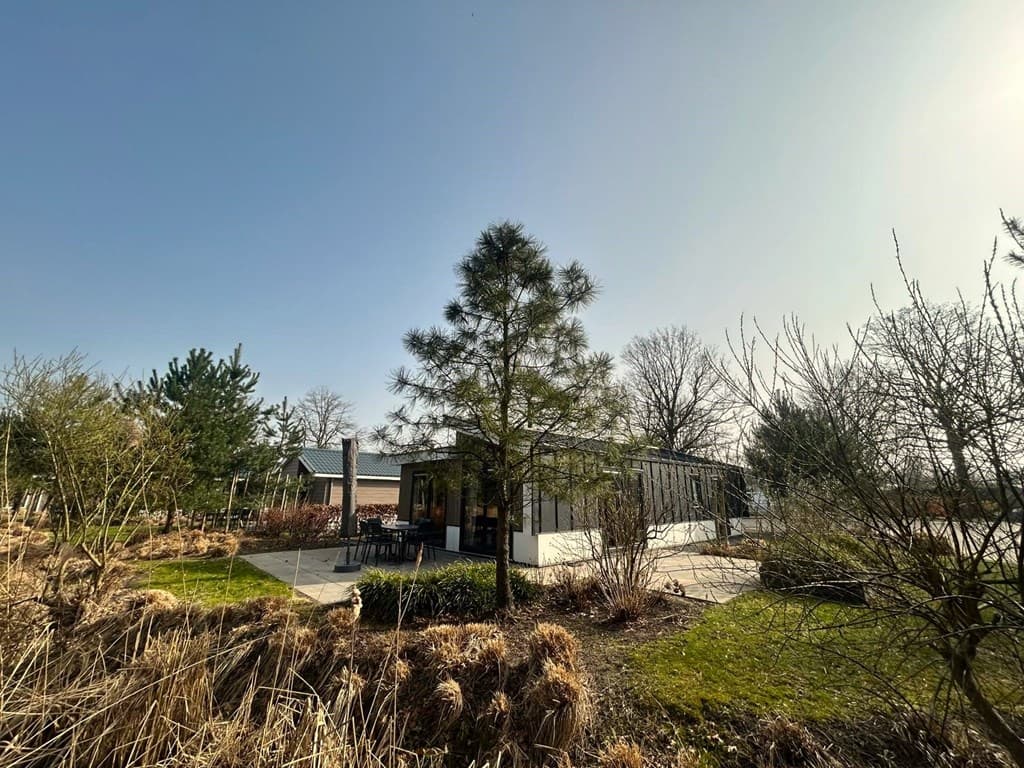 Vakantiewoning omringd door groen, met een terras en een boom in de tuin, onder een heldere blauwe lucht.