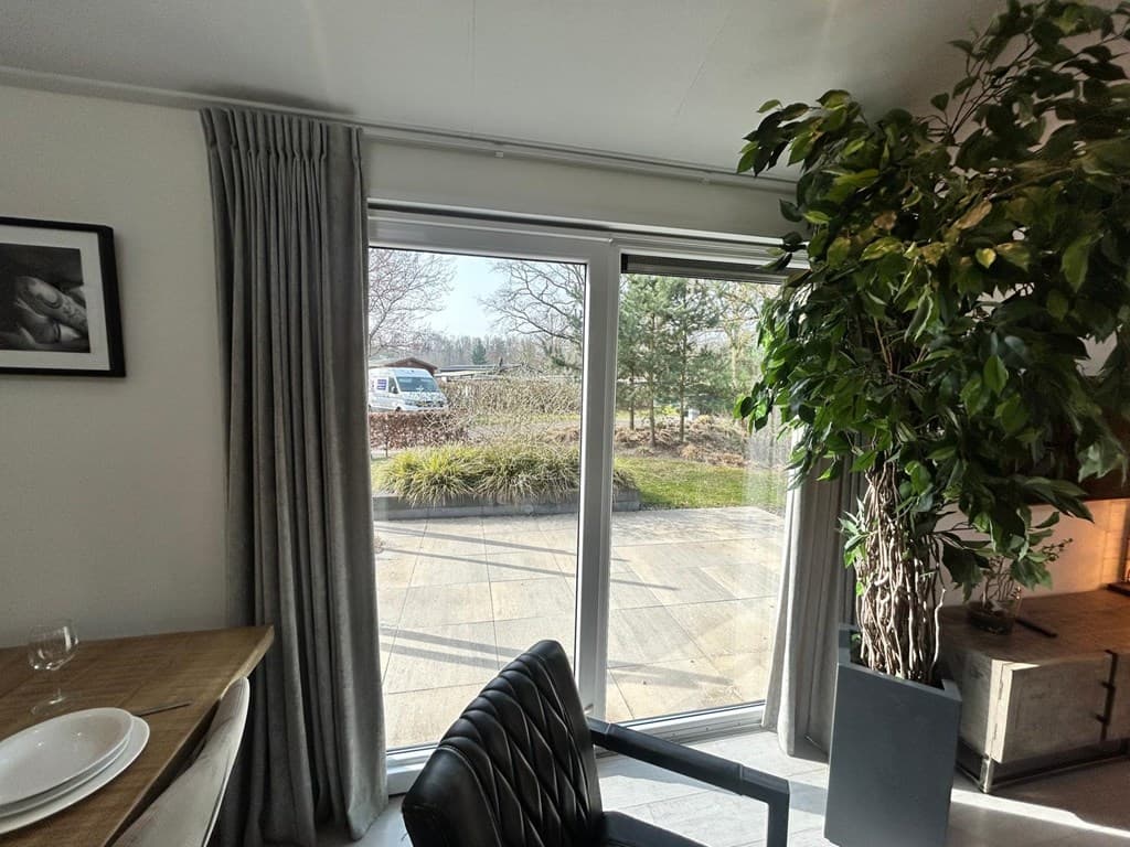 Zicht op een terras met tuin vanuit de woonkamer, groot raam met gordijnen en een plantenbak in de hoek