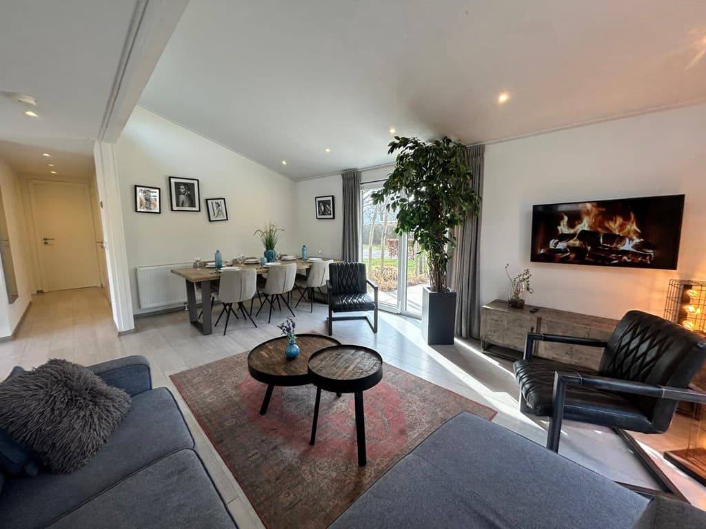 Woonkamer met moderne meubels, grote ramen, en uitzicht op buiten, met eetgedeelte en schouw met vuur.