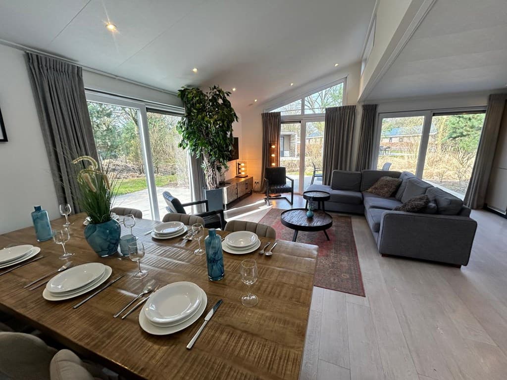 Moderne woonkamer met eettafel en zithoek, grote ramen en een plantenvoorziening aan de wand.