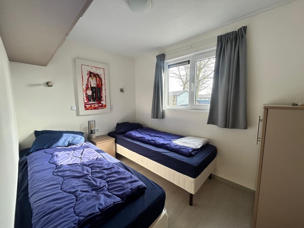 Twee eenpersoonsbedden met blauwe dekbedden en een raam met uitzicht op de buitenruimte in een lichte slaapkamer