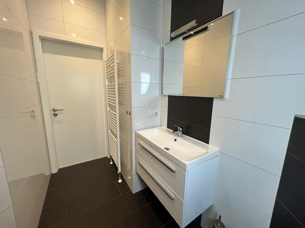 Lichte badkamer met modern wastafelmeubel, spiegel en handdoekenradiator tegen een betegelde wand.