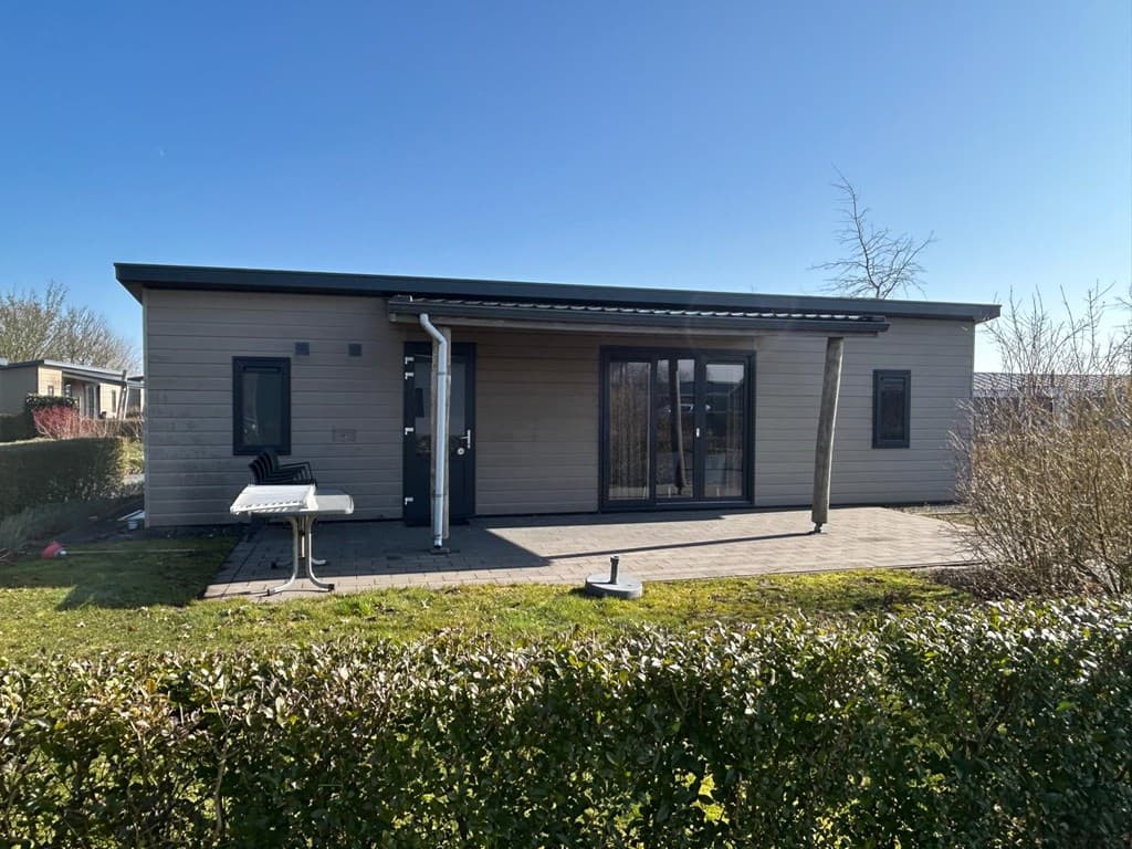 Moderne vakantiewoning met lichte gevel, een betegeld terras en begroeiing aan de voorzijde.