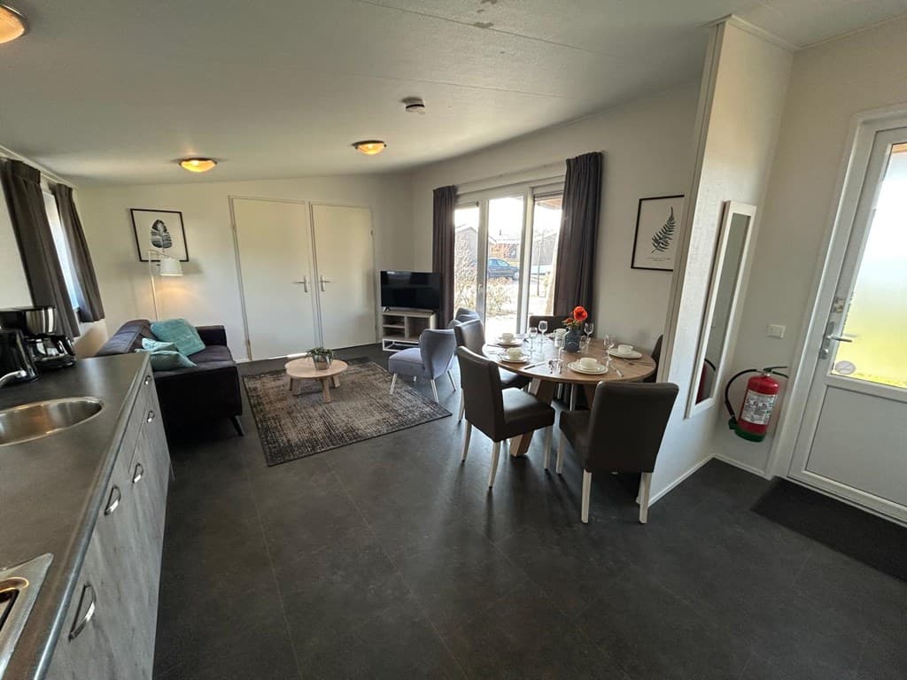 Gezellige woonkamer met eettafel, banken, tv en toegang tot buiten via de deur.