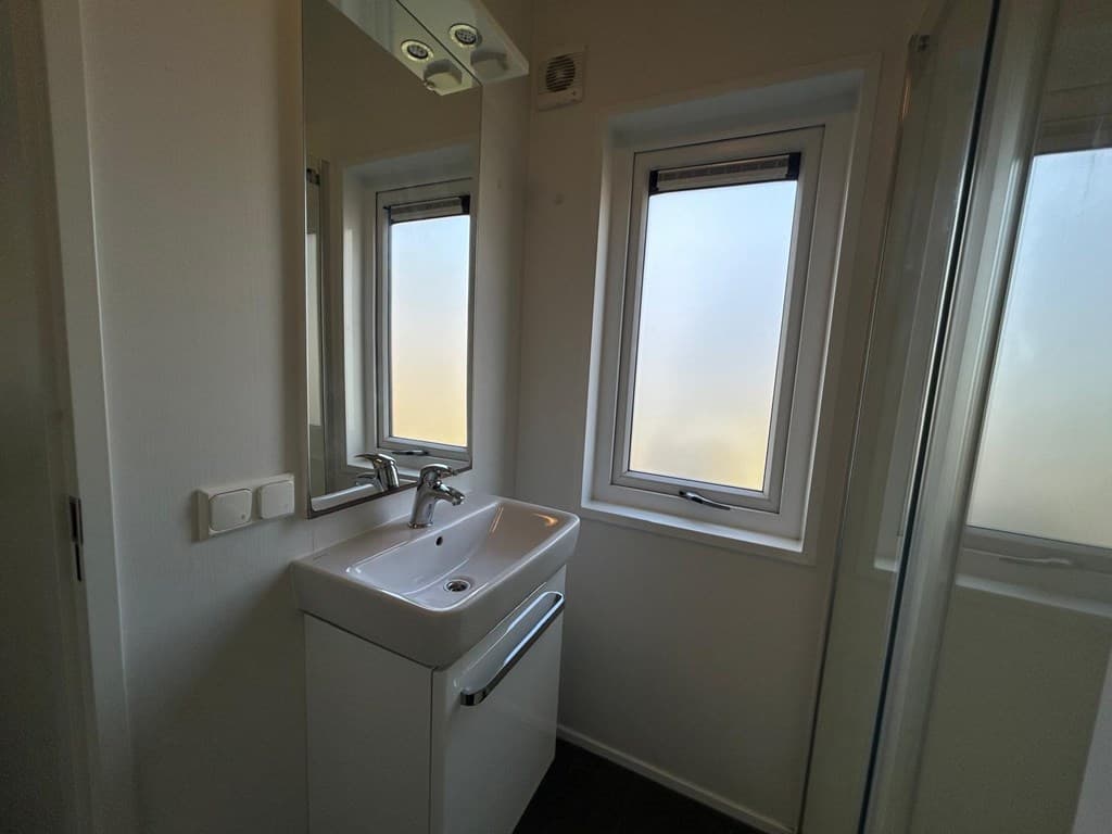 Badkamer met wastafel, spiegel en twee ramen met matglas. Douche zichtbaar in de hoek.