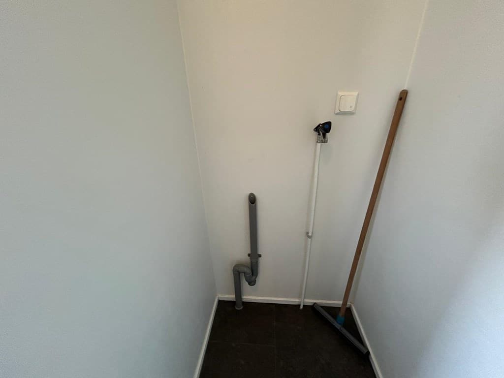 Kleine, lege ruimte met een grijze afvoerbuiging en een bezem in de hoek op een donkere vloer.