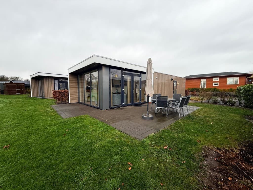 Moderne vakantiewoning met glazen gevel, terras en loungebanken, omgeven door gras en bomen.