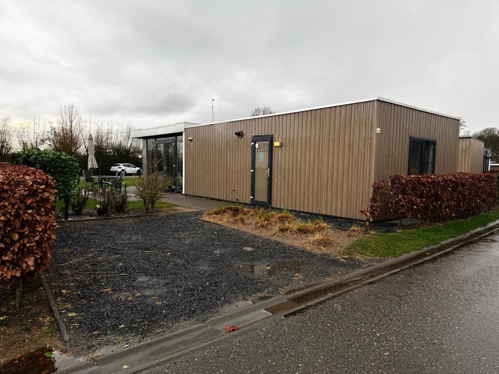Moderne vakantiewoning met houten gevel en voortuin, gelegen langs een verhard pad met groen onderhoud rondom.