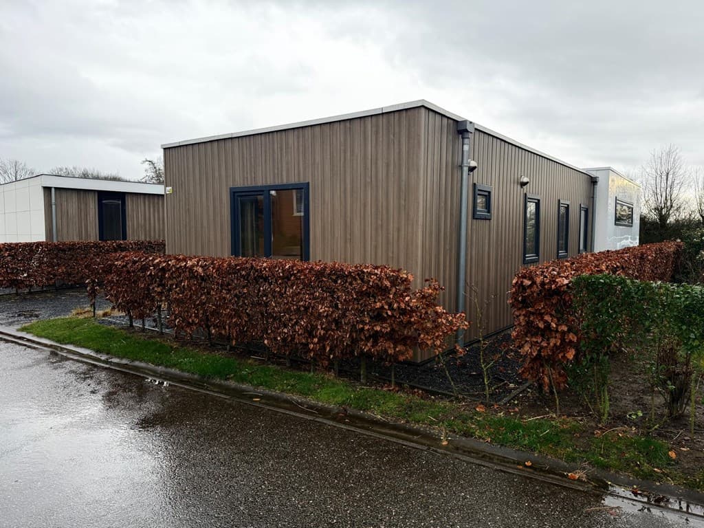 Moderne vakantiewoning met houten gevel, omringd door lage hagen en gelegen aan een verhard pad met natte ondergrond.