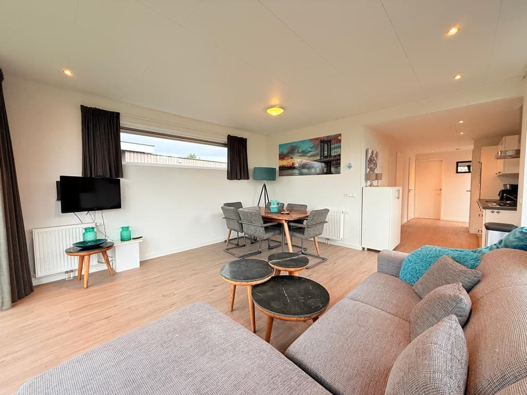 Lichte woonkamer met zithoek, eettafel en keuken, grote ramen en moderne inrichting.