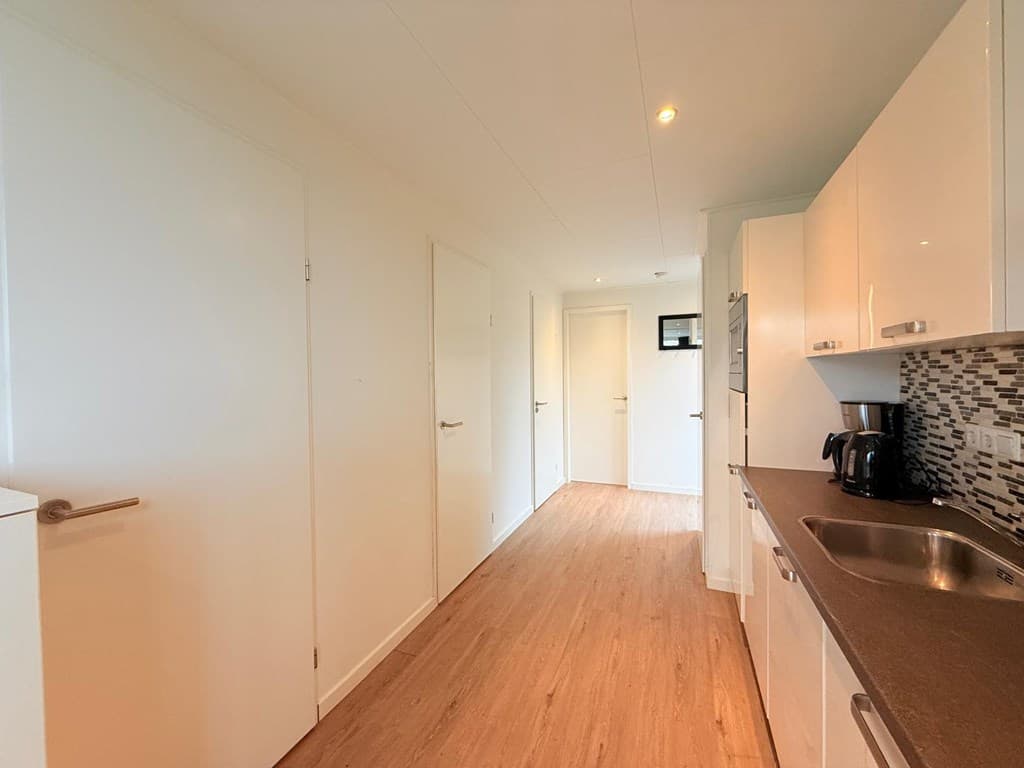 Lichte gang met houten vloer en witte wanden, uitzicht op meerdere deuren en een keuken in moderne stijl.