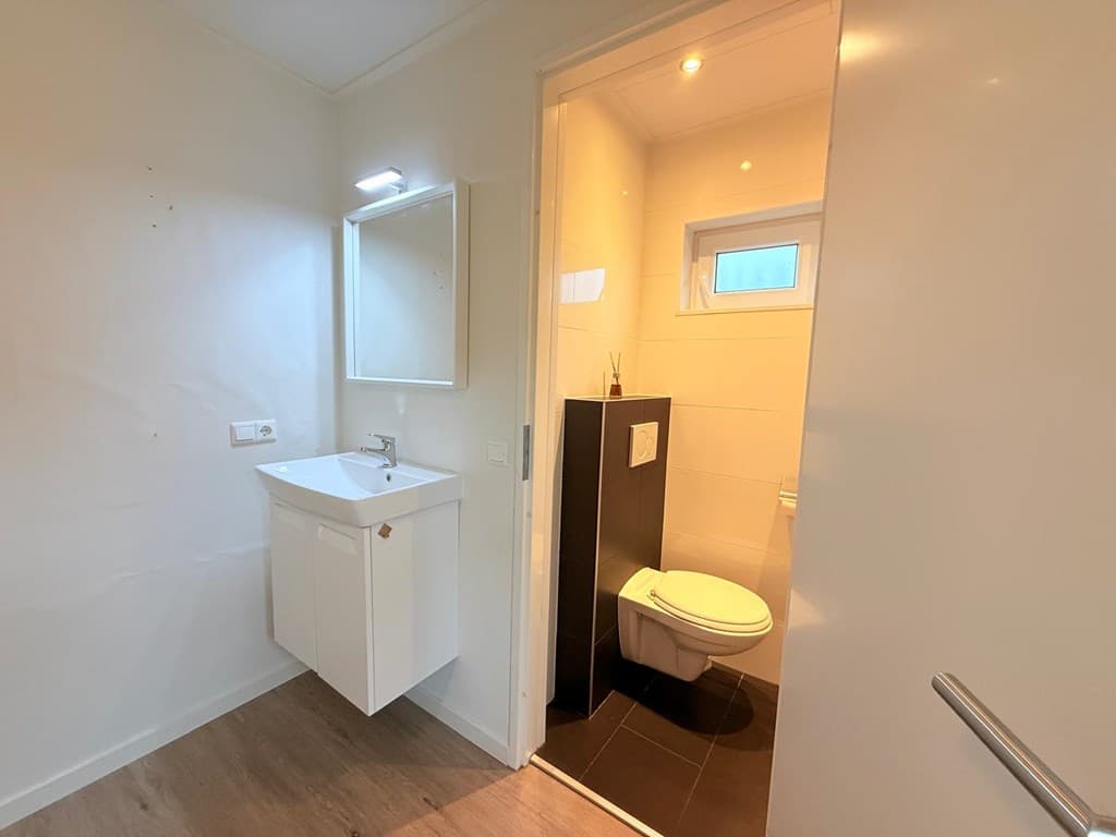 Wit betegeld badkamer met wastafel, spiegel en toilet, lichte muren en een klein raam voor natuurlijk licht