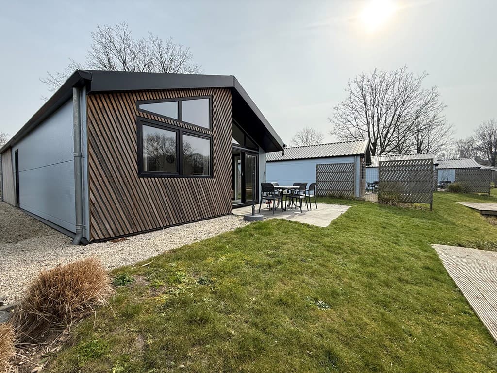 Moderne vakantiewoning met een groen grasveld en terrastafel, omringd door bomen en andere woningen.