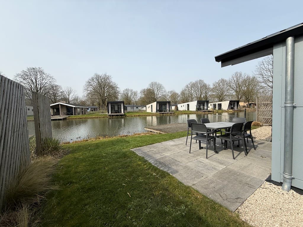 Zitgedeelte met tuinmeubilair aan het water, omringd door vakantiewoningen en bomen in de verte.