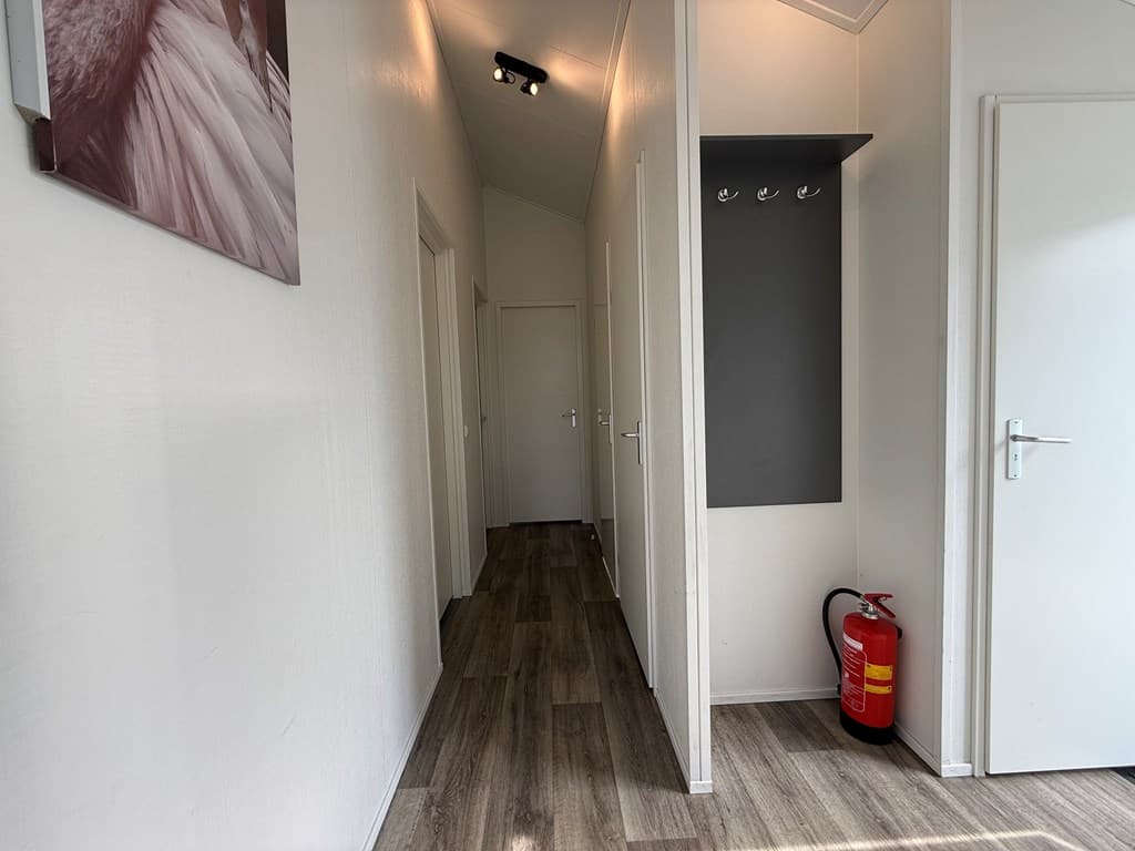 Ingangsruimte met lichte wanden, houten vloer, aan de muur haken en een brandblusser in de hoek.