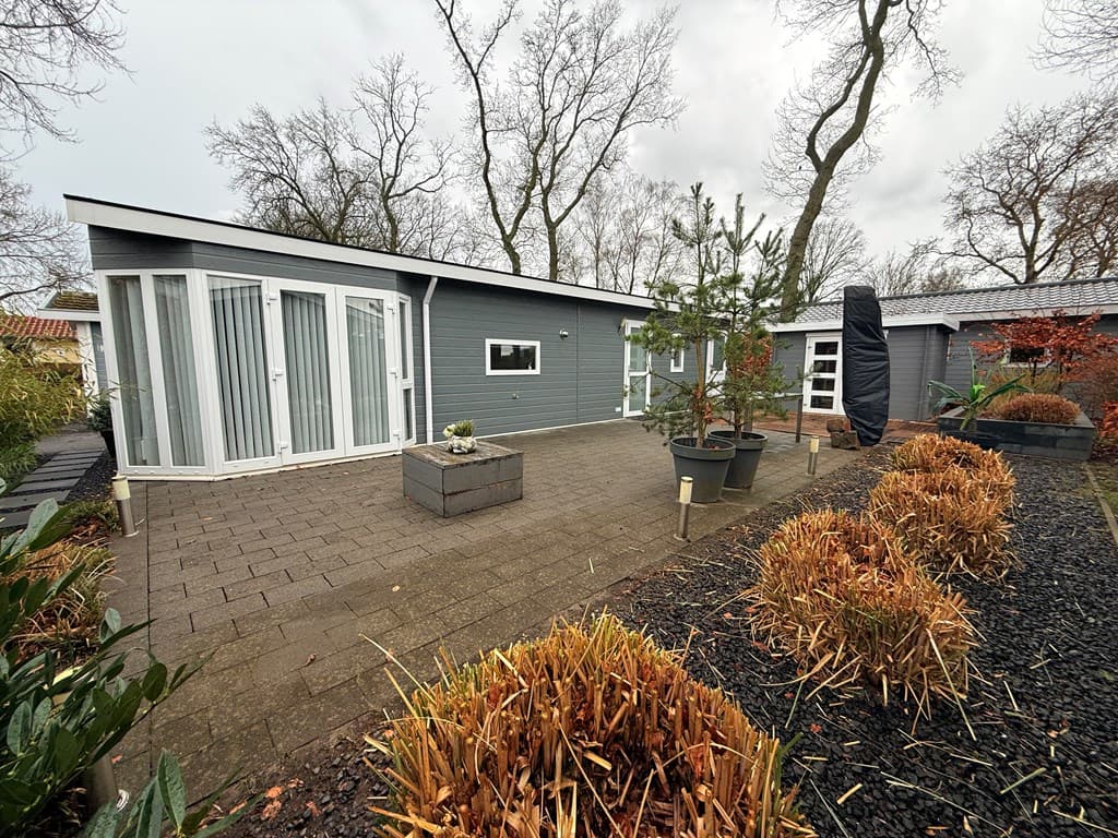 Moderne vakantiewoning met een betegelde patio, plantenbedden en een boom in een bosrijke omgeving.