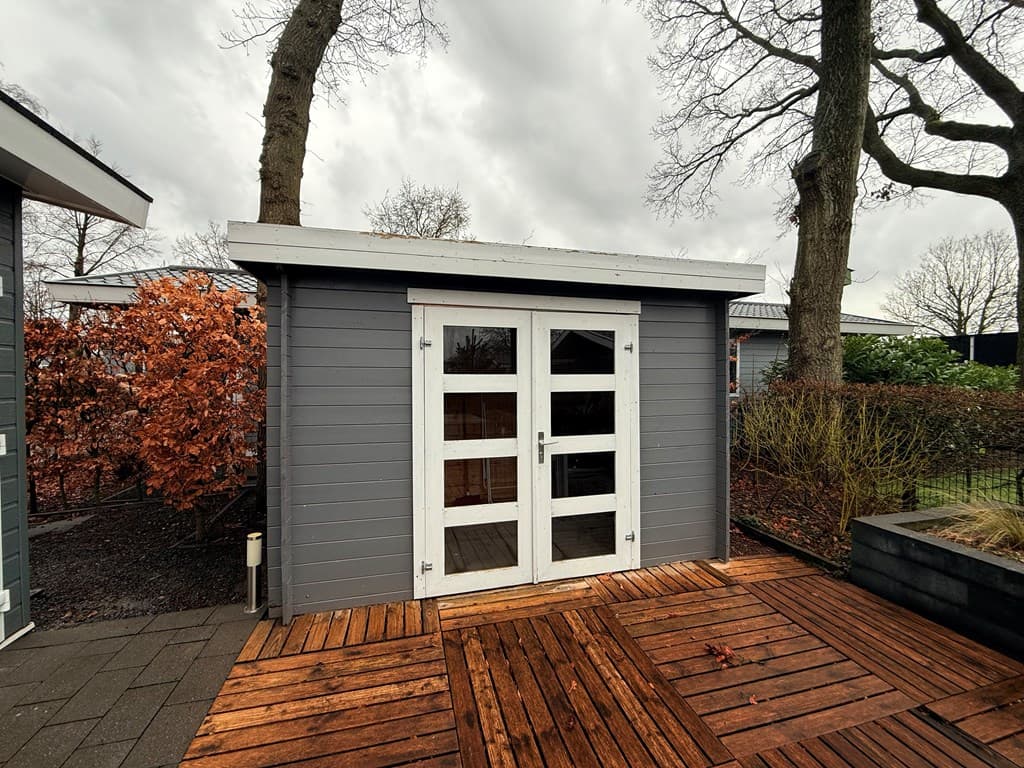 Houten schuur met dubbele deuren op een houten terras, omringd door bomen en struiken