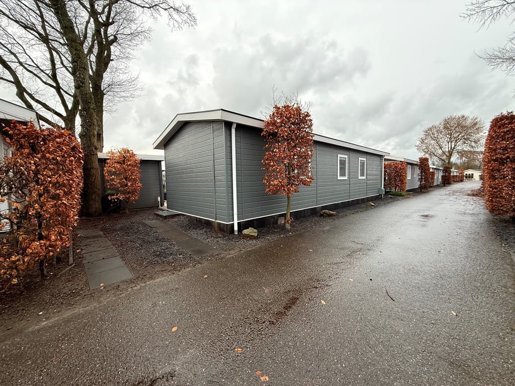Grijze vakantiewoning omringd door hagen met herfstbladeren en een grindpad, onder bewolkte lucht.
