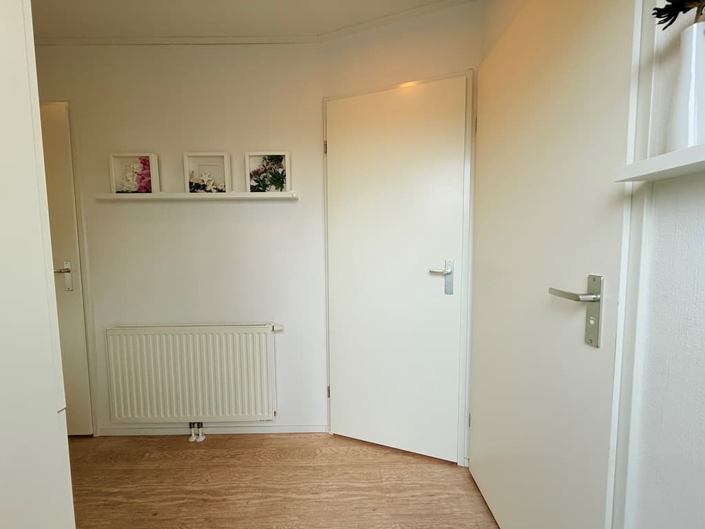 In een lichte gang met laminaatvloer, witte muren en een radiator, drie ingelijste foto's aan de wand.