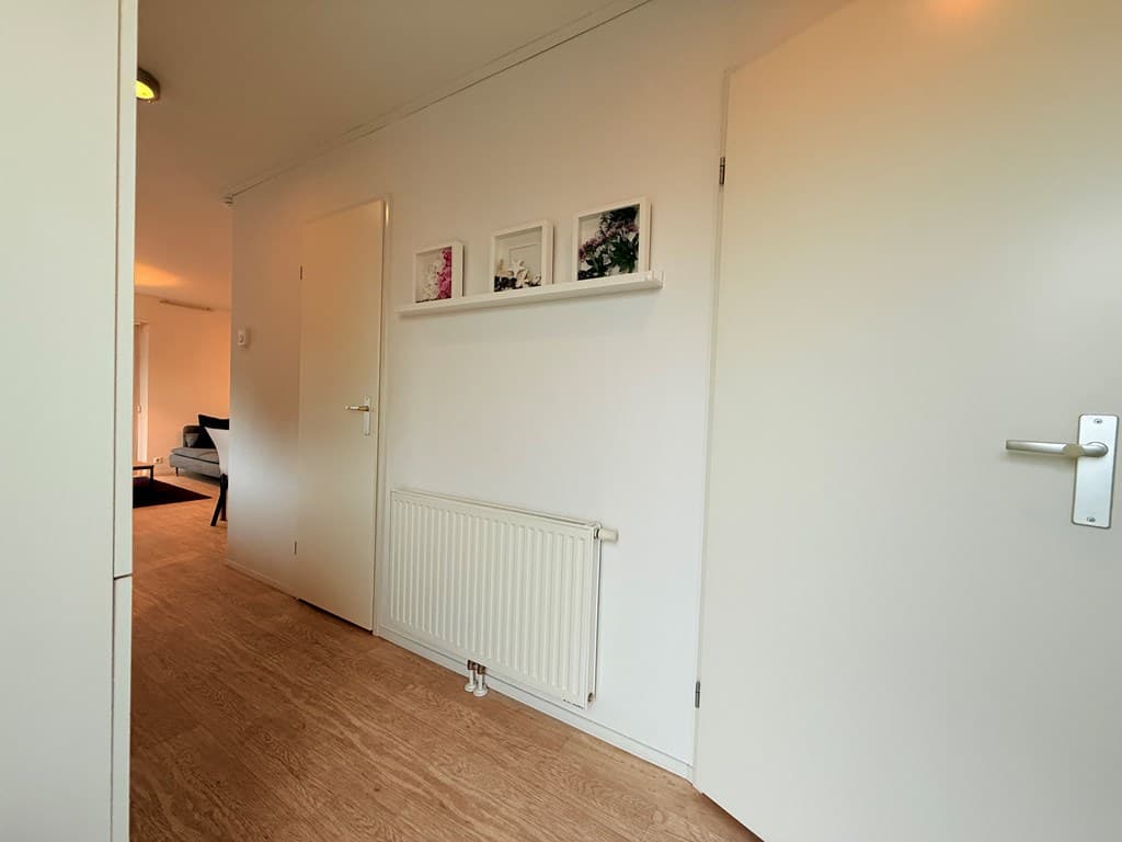 Gang met lichtgekleurde muren, een radiator en een plankenplank met fotolijsten, leidt naar een woonkamer.