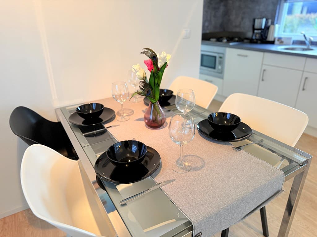 Eetkamertafel met zwarte borden, glazen en een boeket bloemen op een tafelkleed, keuken op de achtergrond zichtbaar