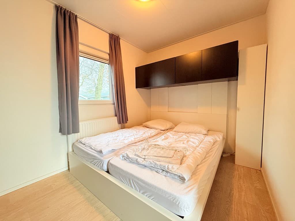 Slaapkamer met een tweepersoonsbed, witte muren en een raam met gordijnen en een kast tegen de muur.