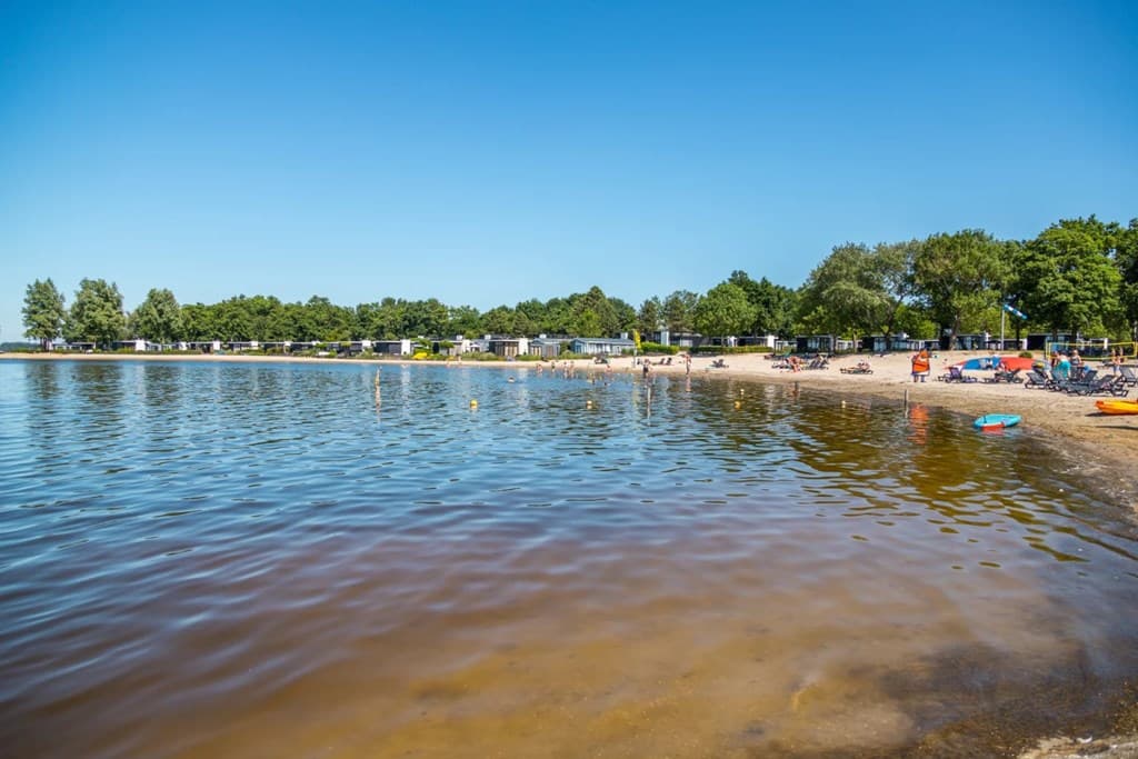 Zicht op het zandstrand en het water bij EuroParcs Bad Hoophuizen, omgeven door bomen en vakantiewoningen