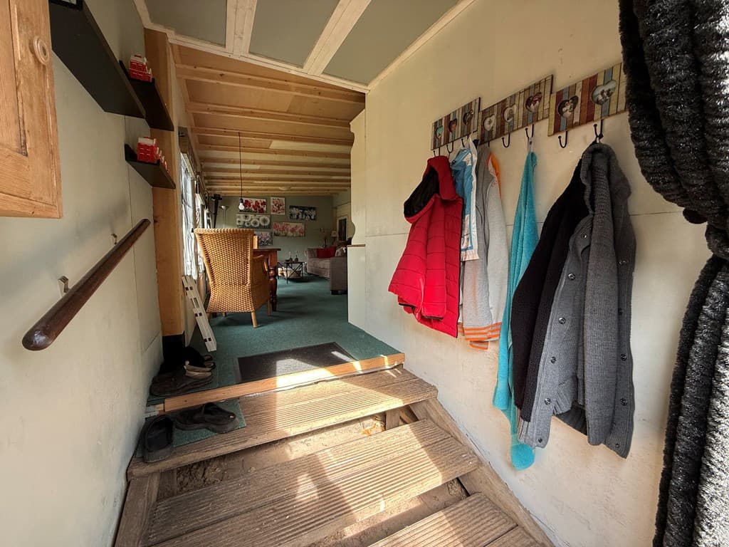 Entree van de woning met jassen aan haken, houten vloer en zicht op een lichte woonkamer met meubels en decoratie